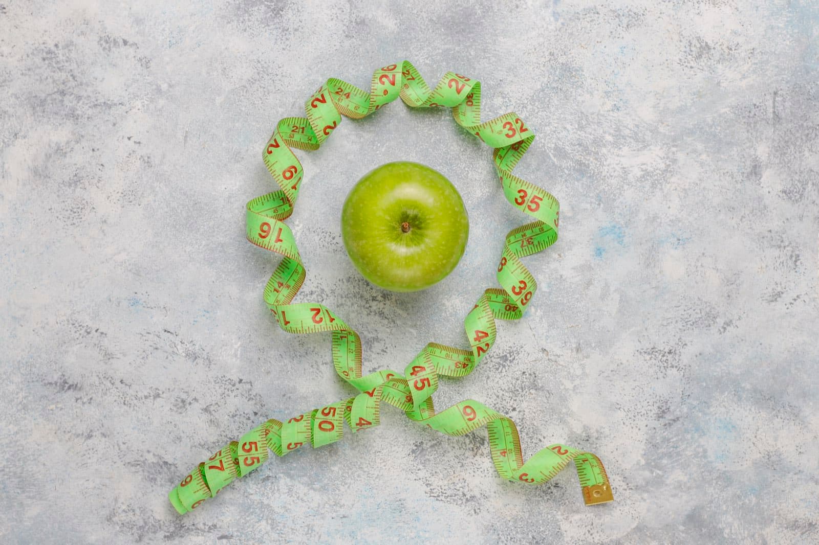 Manzana verde rodeada por una cinta métrica sobre una superficie de concreto claro