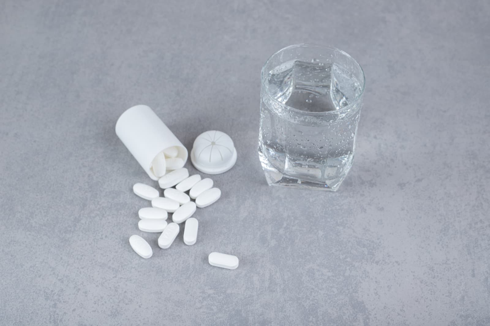Pastillas blancas junto a un frasco abierto y un vaso de agua sobre una superficie gris